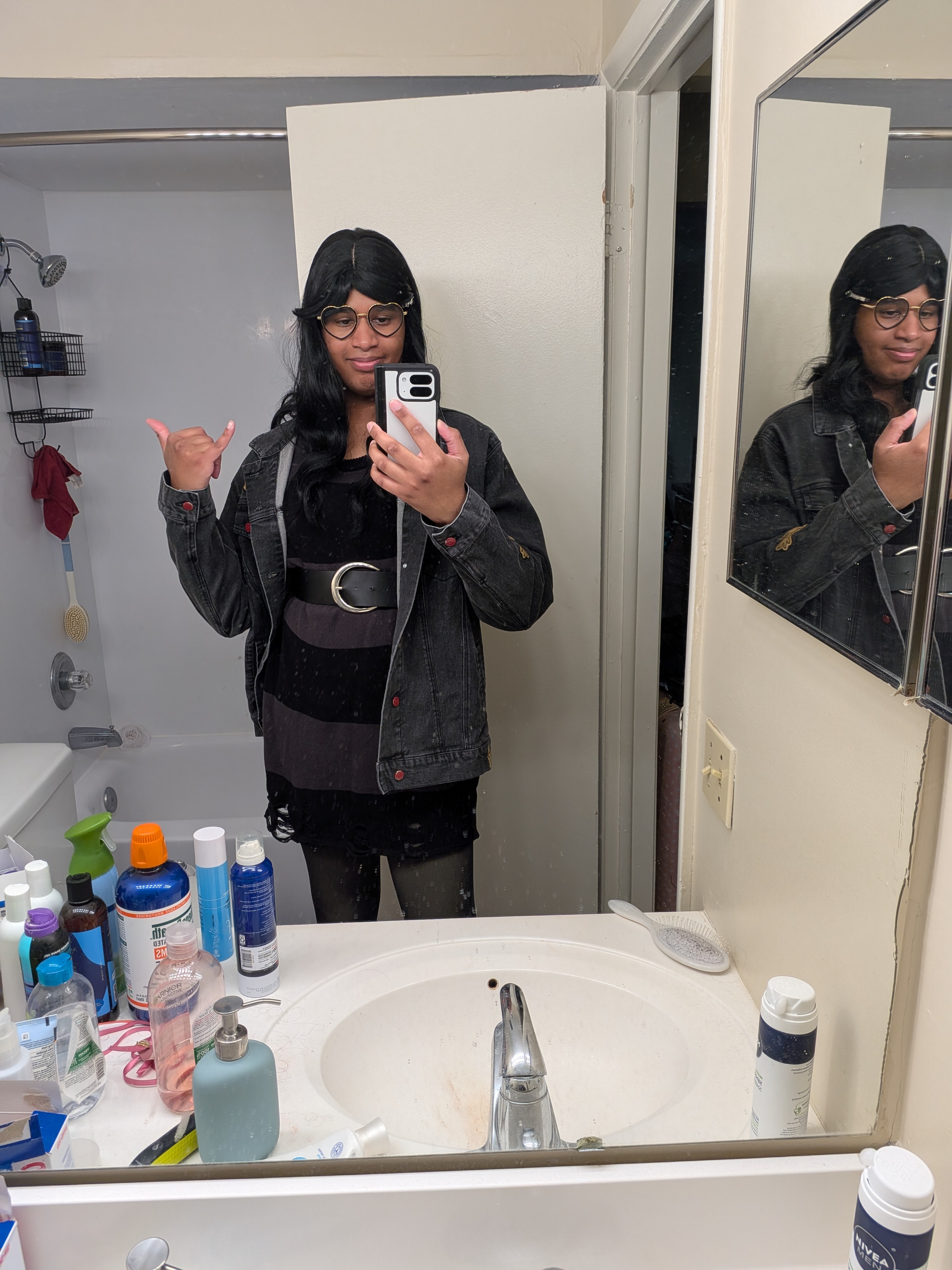 Adrianna wearing a long black wig with heart glasses. I have on a tunic that is dark green as the base with larger black stripes down and frayed hem on the bottom. Over it I'm wearing the Pokemon Worlds 2026 Jean Jacket. Bottoms are black tights.