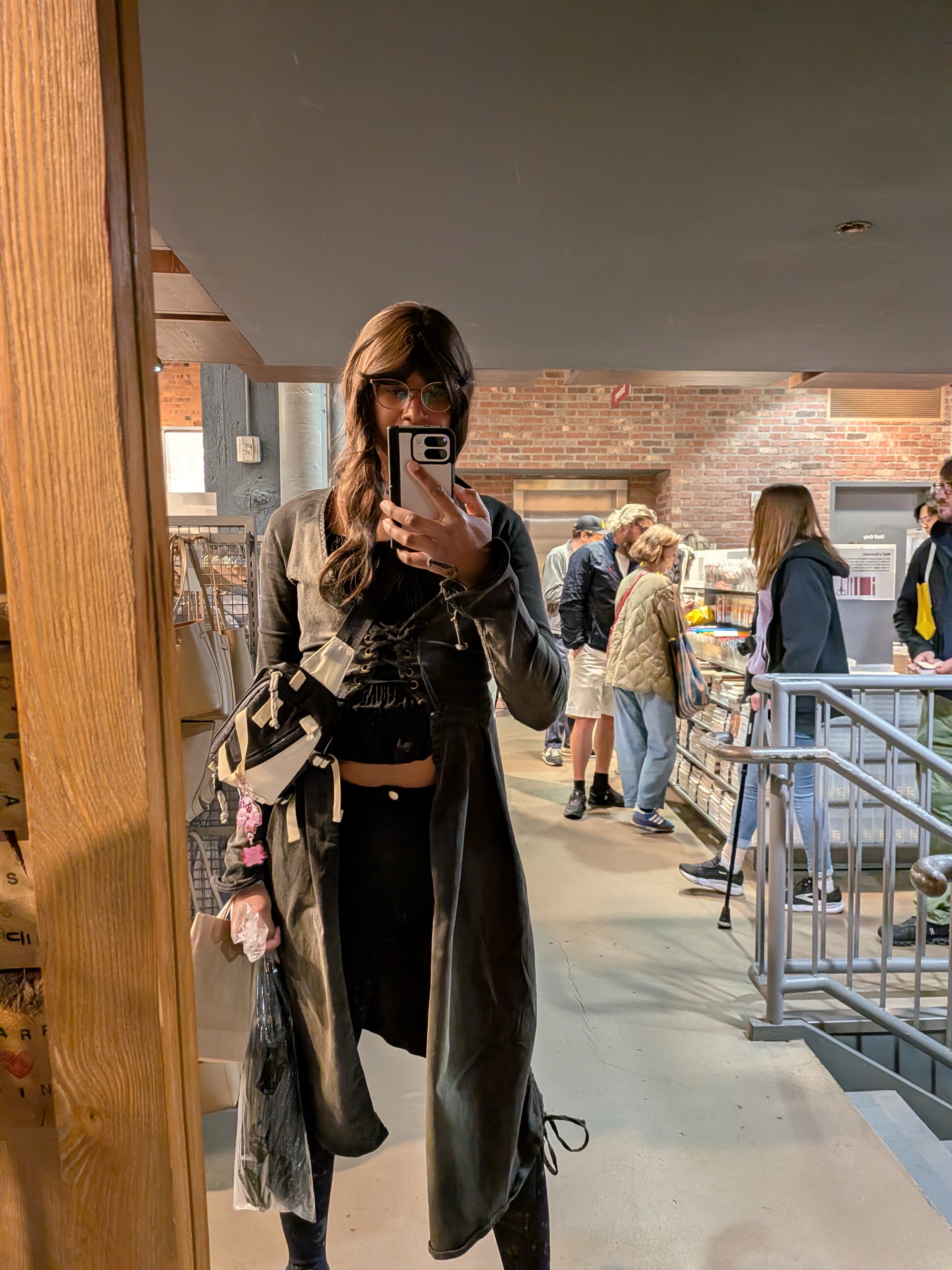 Adrianna wearing a long brown wig with blonde highlights and catseye glasses. I have on a dark grey corset duster coat. Underneath I have on a black cropped blouse. I have on black shorts over starry tights.