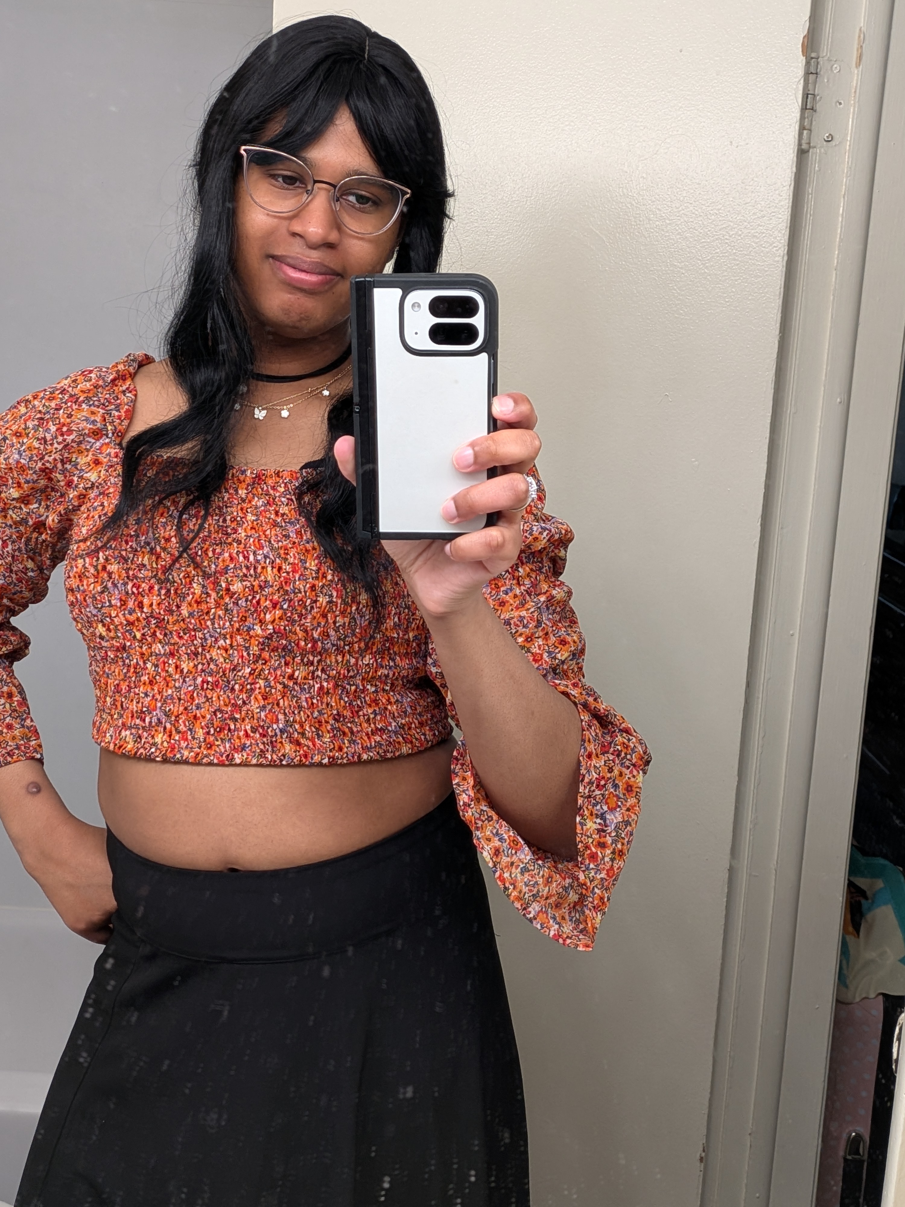 Adrianna wearing a long black wig with catseye glasses. An orange floral printed Crop top and black maxi skirt
