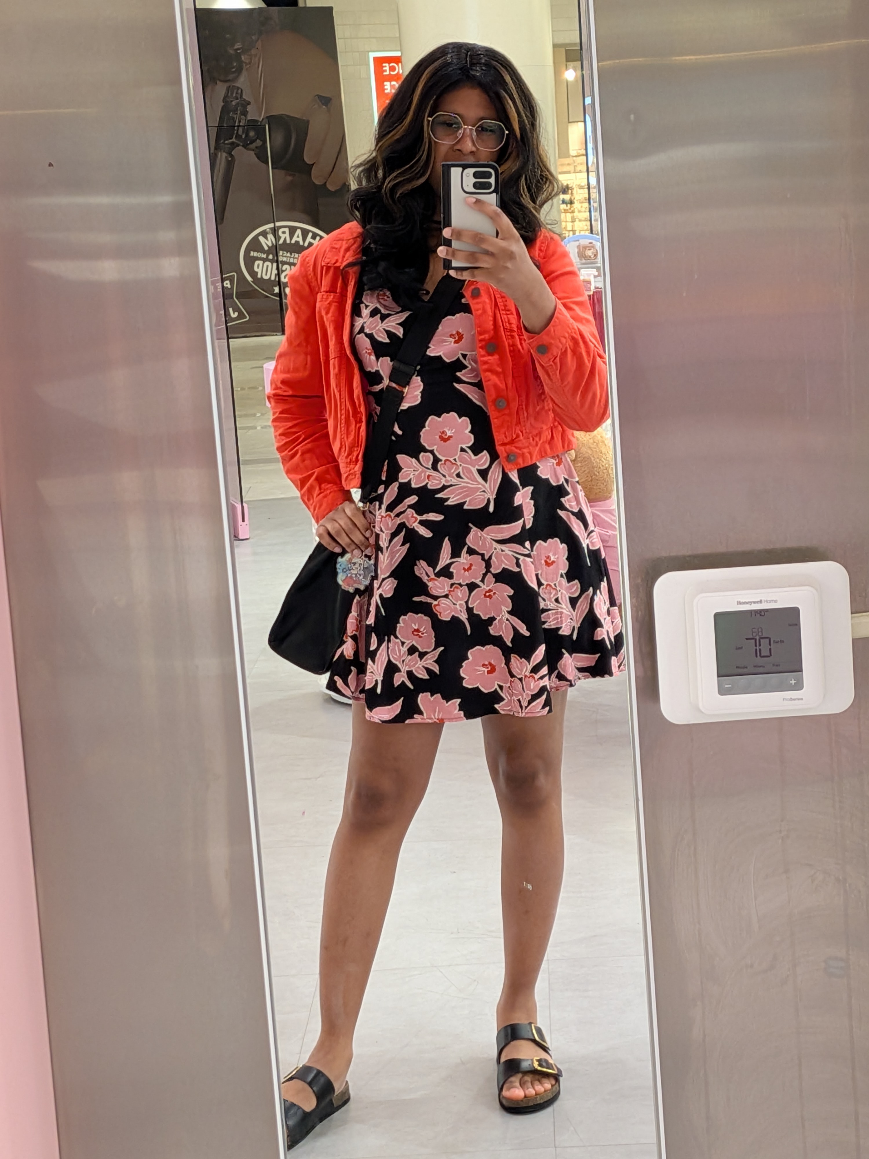 Adrianna wearing a long wavy brown wig with blonde highlights and geometric-octogonal glasses. A short black dress with pink floral prints under a red jean jacket